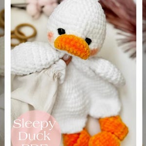 May include: A handmade white crochet duck toy with an orange beak and feet. The duck has black eyes and rosy cheeks. The text "Sleepy Duck PDF" is on a pink circle in the lower left corner. The toy is soft and cuddly.