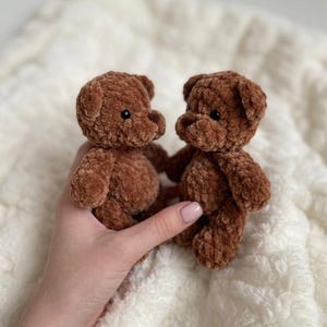 May include: Two small, handmade brown teddy bears crafted from soft, plush chenille yarn. Each bear features a rounded head, small ears, and dark, round plastic eyes. The texture of the yarn is thick and fuzzy, giving the bears a tactile appearance.