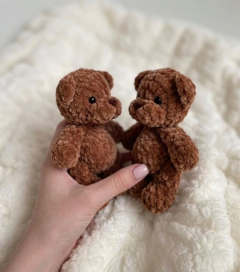 May include: Two small, handmade brown teddy bears crafted from soft, plush chenille yarn. Each bear features a rounded head, small ears, and dark, round plastic eyes. The texture of the yarn is thick and fuzzy, giving the bears a tactile appearance.