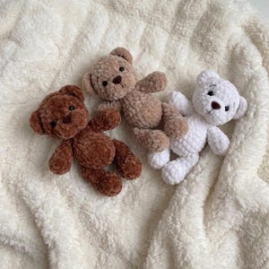 May include: Three small plush teddy bears made of soft, textured yarn. One bear is dark brown, one is light tan, and one is white. Each bear features round ears, small dark eyes, and a stitched brown nose, arranged together on a white fleece fabric.