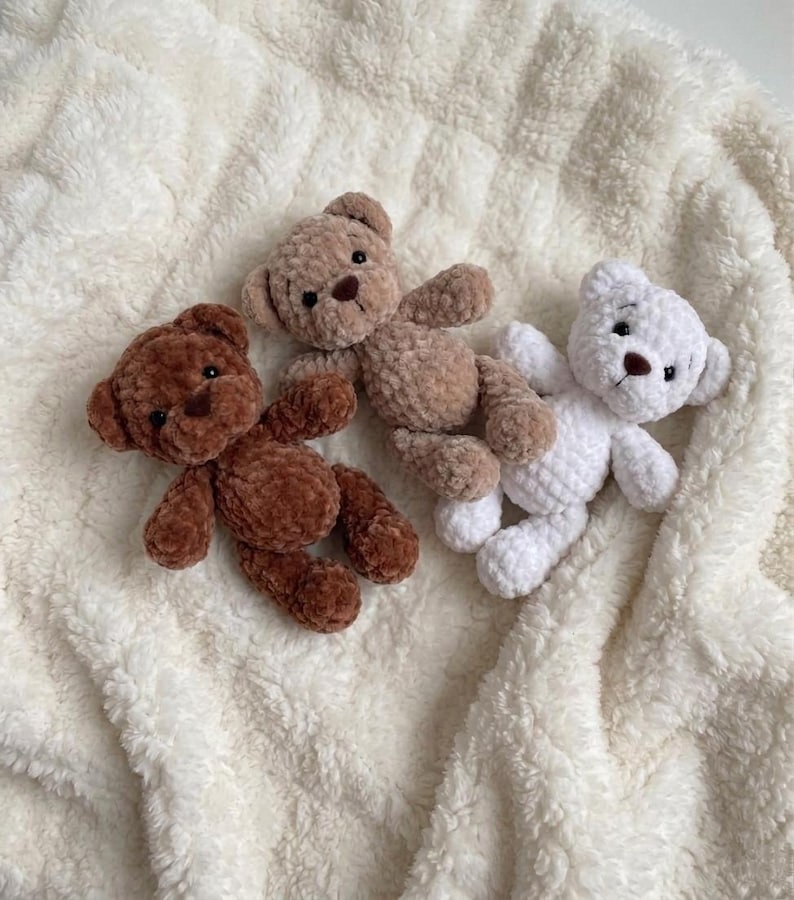 May include: Three small plush teddy bears made of soft, textured yarn. One bear is dark brown, one is light tan, and one is white. Each bear features round ears, small dark eyes, and a stitched brown nose, arranged together on a white fleece fabric.