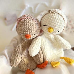 Könnte beinhalten: Zwei gehäkelte Entenküken, eines beige und eines weiß, kuscheln eng aneinander. Das beige Küken hat eine rosa Schleife, das weiße eine beige Schleife. Beide haben orangefarbene Schnäbel und Füße und geschlossene Augen. Ein gemütliches, handgefertigtes Produkt.