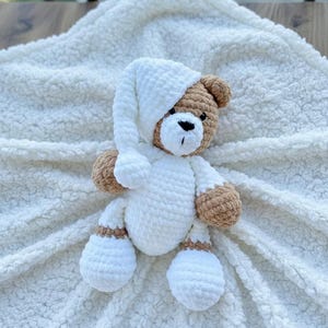 May include: A handmade crochet teddy bear wearing a white nightcap and pajamas, with brown accents on the head, arms, and legs. The bear is lying on a soft, white, textured blanket, creating a cozy and comforting scene.