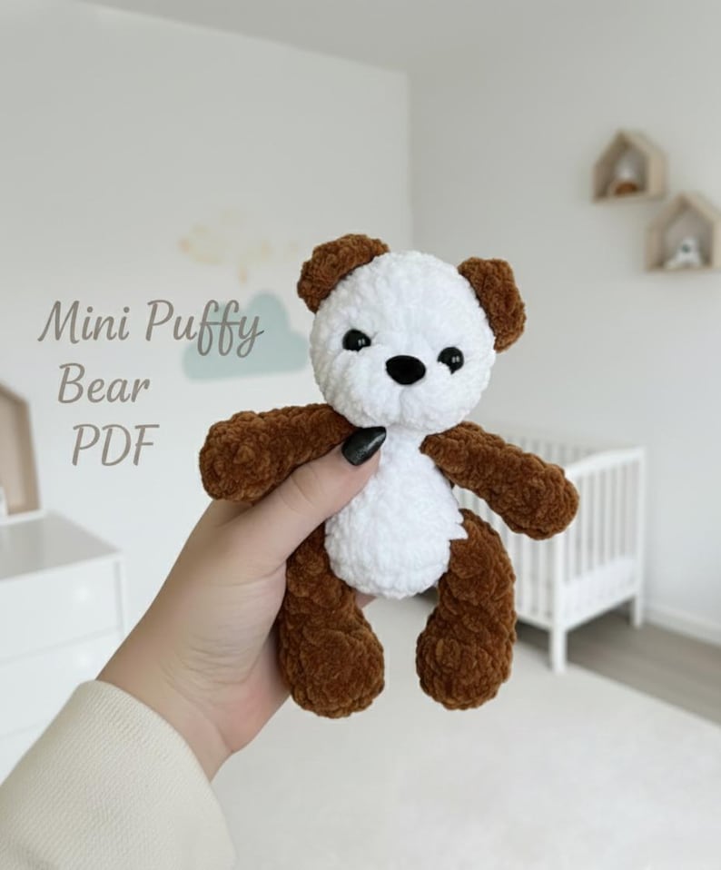 May include: A small, plush, crocheted bear toy featuring a white head and torso with brown ears, arms, and legs. The bear has black plastic eyes and a black nose. The text 'Mini Puffy Bear PDF' is visible on the left side of the image.