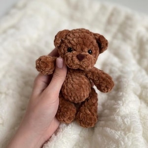 May include: A small, brown, plush teddy bear crafted from soft, textured yarn. The toy features rounded ears, a dark brown stitched nose, and small black bead eyes. The bear has a simple, compact design with articulated limbs and a fuzzy finish.