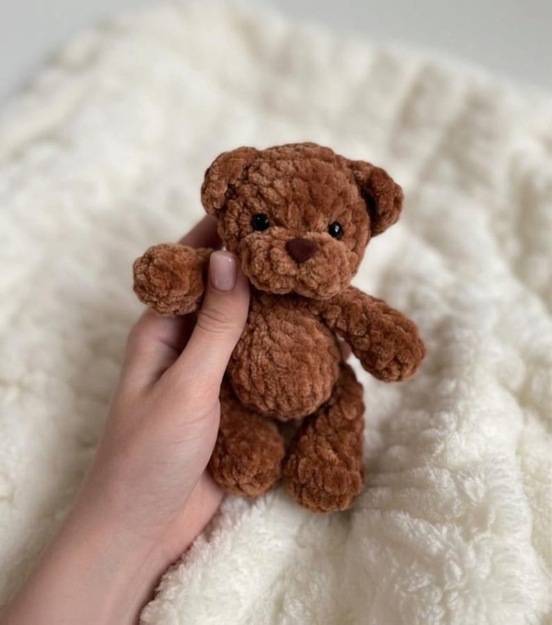 May include: A small, brown, plush teddy bear crafted from soft, textured yarn. The toy features rounded ears, a dark brown stitched nose, and small black bead eyes. The bear has a simple, compact design with articulated limbs and a fuzzy finish.