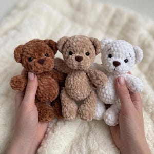 May include: Three small plush teddy bears made of soft, textured yarn. The bears are arranged in a row, featuring a dark brown bear on the left, a light tan bear in the centre, and a white bear on the right. Each bear has black eyes and a nose.
