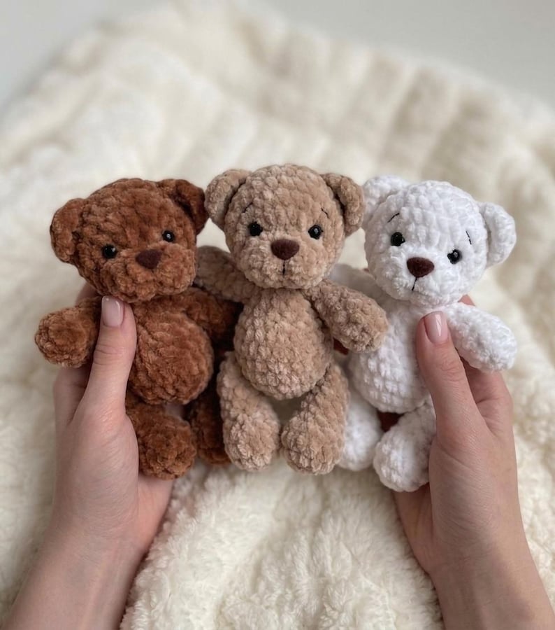 May include: Three small plush teddy bears made of soft, textured yarn. The bears are arranged in a row, featuring a dark brown bear on the left, a light tan bear in the centre, and a white bear on the right. Each bear has black eyes and a nose.