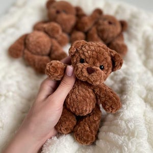 May include: A small, brown, plush teddy bear made of soft, textured yarn. The bear features round ears, a dark brown stitched nose, and small black bead eyes. It has a rounded body with articulated limbs and a fuzzy, tactile surface texture.