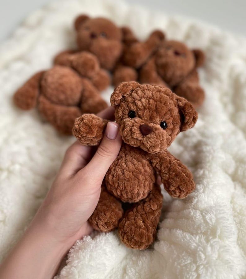 May include: A small, brown, plush teddy bear made of soft, textured yarn. The bear features round ears, a dark brown stitched nose, and small black bead eyes. It has a rounded body with articulated limbs and a fuzzy, tactile surface texture.