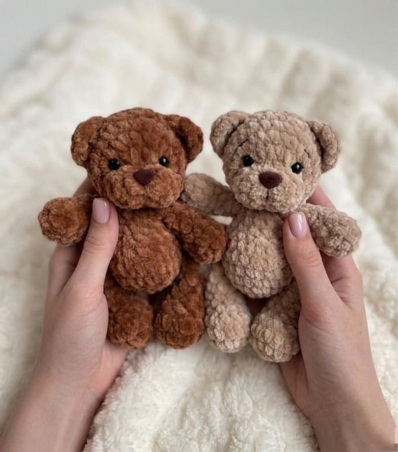 May include: Two small plush teddy bears made of soft, textured yarn. One bear is a deep chocolate brown, and the other is a lighter beige colour. Both bears feature round ears, dark plastic eyes, and a small, dark brown stitched nose on their faces.