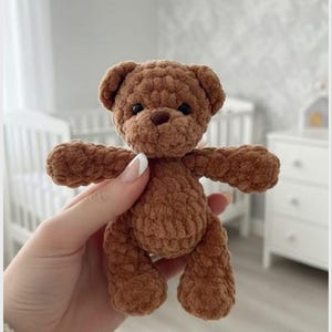 May include: A small, brown, crocheted teddy bear made from soft, plush yarn. The bear features a rounded head with two small ears, a dark brown nose, and two black bead eyes. Its body has short arms and legs, with a textured, fuzzy appearance.