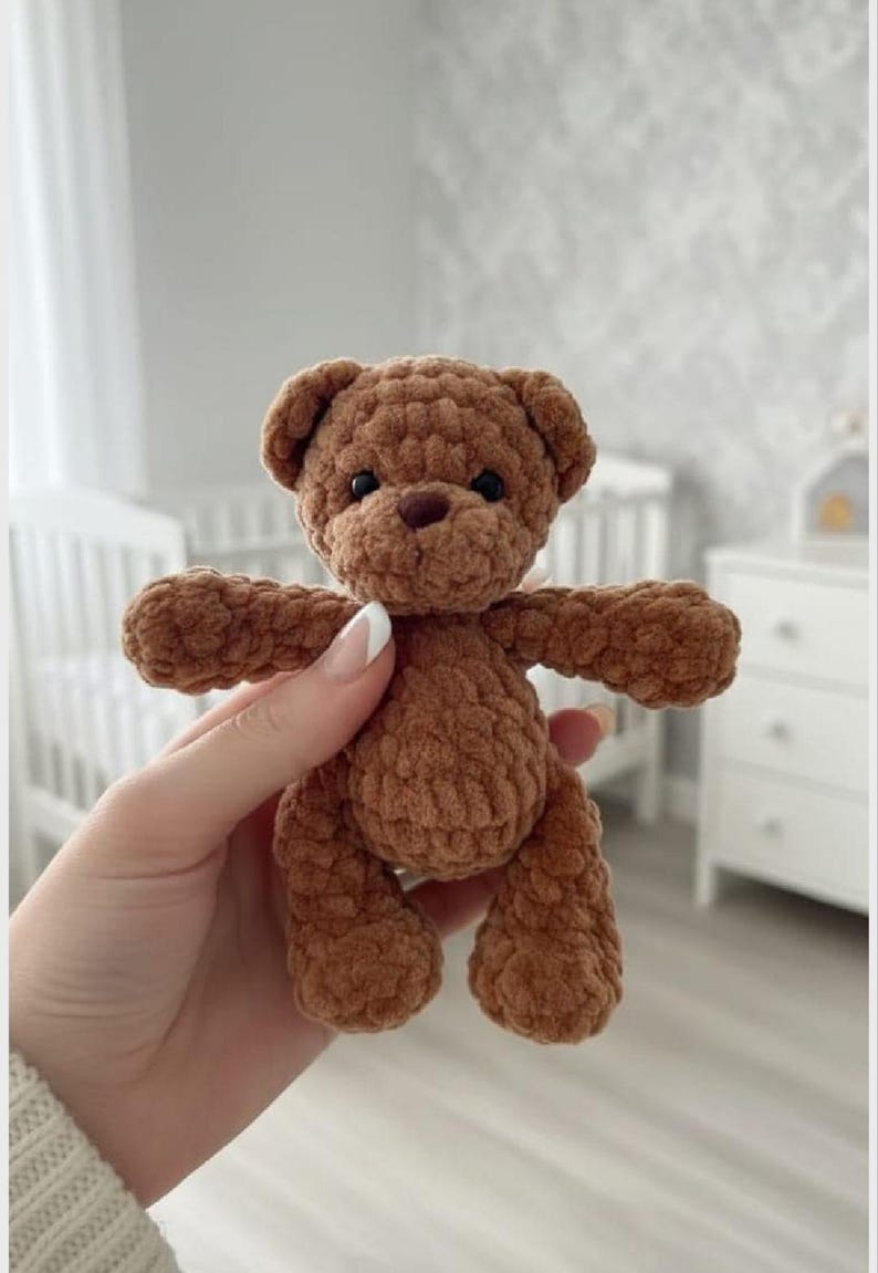 May include: A small, brown, crocheted teddy bear made from soft, plush yarn. The bear features a rounded head with two small ears, a dark brown nose, and two black bead eyes. Its body has short arms and legs, with a textured, fuzzy appearance.