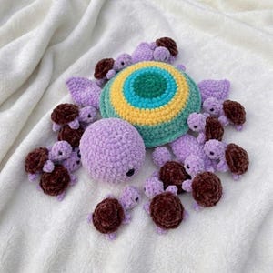 May include: A collection of handmade crochet items, including a large, round, multi-colored turtle shell, a smaller lavender turtle body, and numerous small turtles with brown shells and lavender bodies. The items are arranged on a white, textured surface.