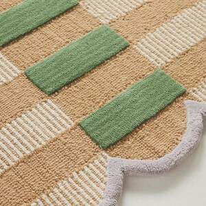 May include: A beige and white checkered rug with green rectangular accents. The rug has a scalloped edge in a light purple color. The rug is made of a soft, textured material.