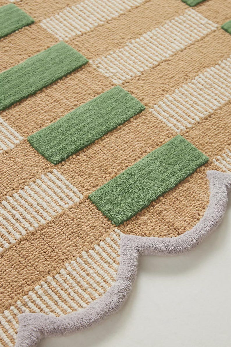 May include: A beige and white checkered rug with green rectangular accents. The rug has a scalloped edge in a light purple color. The rug is made of a soft, textured material.