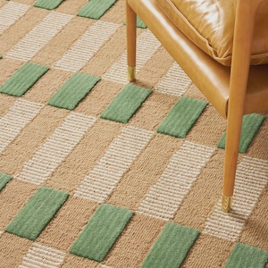 May include: A tan and green patterned rug with a repeating rectangular design. The rug is made of a textured material and has a scalloped edge. A wooden chair with a tan leather cushion sits on the rug.