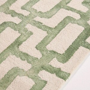 May include: Close-up of a rug with a geometric pattern. The rug has a repeating design of light green shapes against a cream background. The texture appears soft and plush, suggesting a comfortable floor covering.