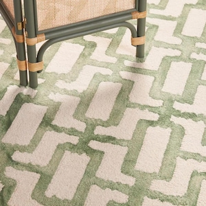 May include: A close-up of a green and cream rug with a geometric pattern. The rug is paired with a green and rattan room divider. The rug's design features a repeating pattern of green shapes against a cream background, creating a textured look.