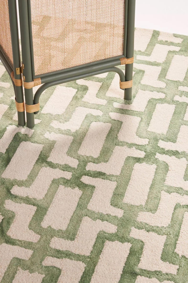 May include: A close-up of a green and cream rug with a geometric pattern. The rug is paired with a green and rattan room divider. The rug's design features a repeating pattern of green shapes against a cream background, creating a textured look.