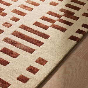 May include: A beige rug with a geometric pattern of rectangular shapes in varying sizes and shades of brown. The rug's design features a repeating pattern of blocks and lines, creating a modern and textured look. The rug is placed on a light-colored wooden floor.