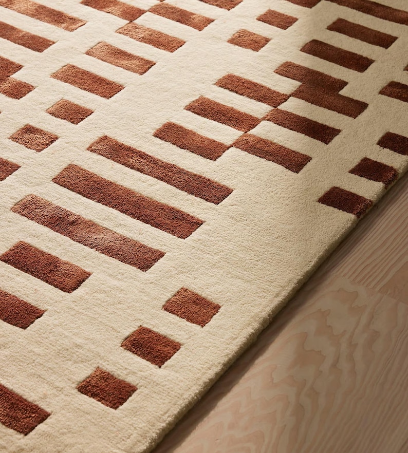 May include: A beige rug with a geometric pattern of rectangular shapes in varying sizes and shades of brown. The rug's design features a repeating pattern of blocks and lines, creating a modern and textured look. The rug is placed on a light-colored wooden floor.