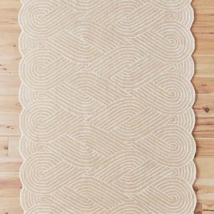 May include: A beige rug with a woven design. The rug features a repeating pattern of intertwined loops and a scalloped edge. The neutral color and textured surface create a cozy aesthetic.