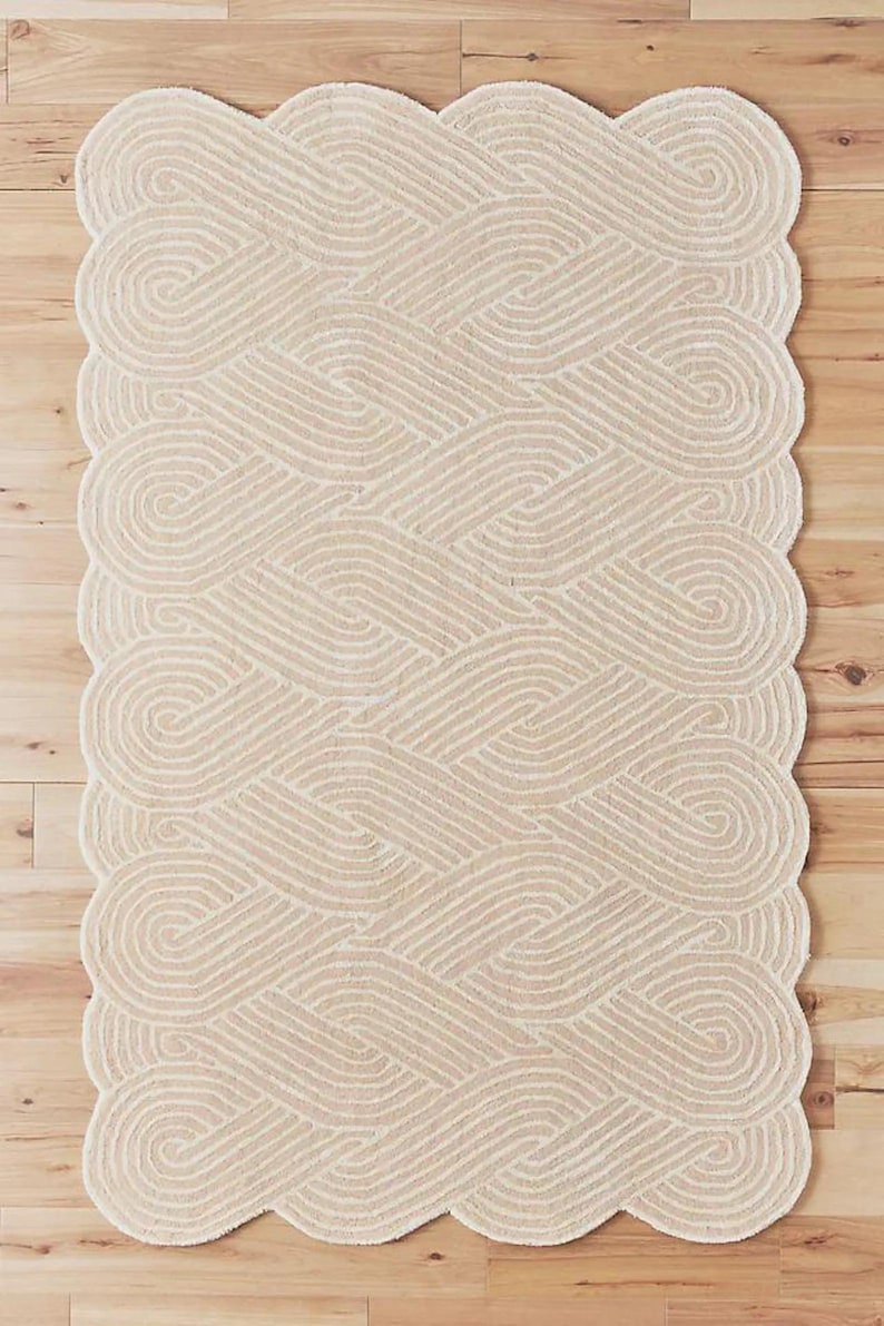 May include: A beige rug with a woven design. The rug features a repeating pattern of intertwined loops and a scalloped edge. The neutral color and textured surface create a cozy aesthetic.