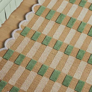 May include: A decorative rug with a repeating pattern of tan, white, and green rectangles. The rug has a scalloped edge with a gray border. The rug is placed on a wooden floor.