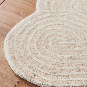May include: A cream-colored, circular rug with a spiral design. The rug has a soft, textured surface and a light beige color. The rug is placed on a wooden floor.