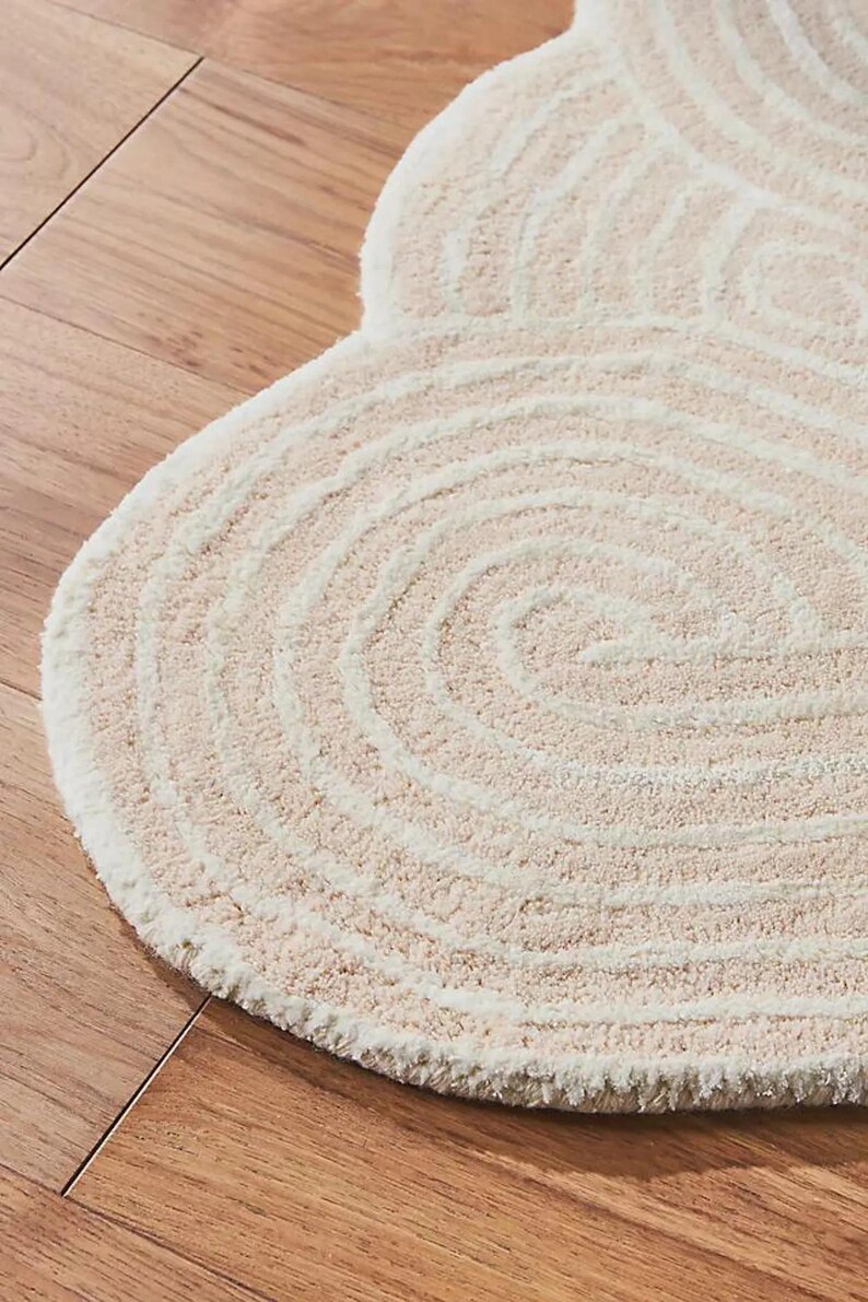 May include: A cream-colored, circular rug with a spiral design. The rug has a soft, textured surface and a light beige color. The rug is placed on a wooden floor.
