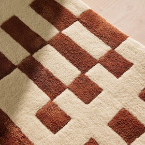 May include: Close-up of a textured rug with a geometric pattern. The rug features alternating rectangular blocks of cream and brown. The brown blocks are a darker shade, creating a contrast against the lighter cream background. The rug appears soft and plush.