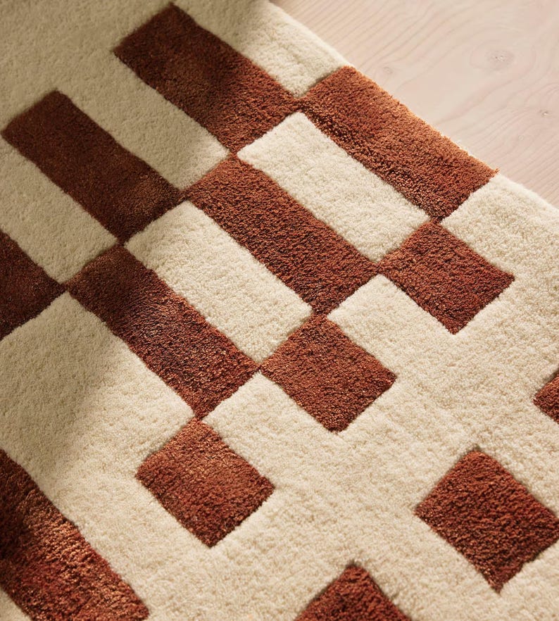 May include: Close-up of a textured rug with a geometric pattern. The rug features alternating rectangular blocks of cream and brown. The brown blocks are a darker shade, creating a contrast against the lighter cream background. The rug appears soft and plush.