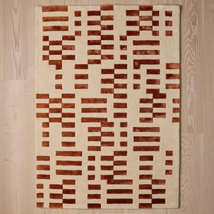 May include: Rectangular rug with a cream-colored background and a repeating pattern of rust-colored horizontal and vertical rectangular blocks. The rug's design is geometric and modern, suitable for a living room or bedroom.