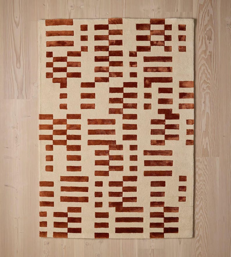 May include: Rectangular rug with a cream-colored background and a repeating pattern of rust-colored horizontal and vertical rectangular blocks. The rug's design is geometric and modern, suitable for a living room or bedroom.
