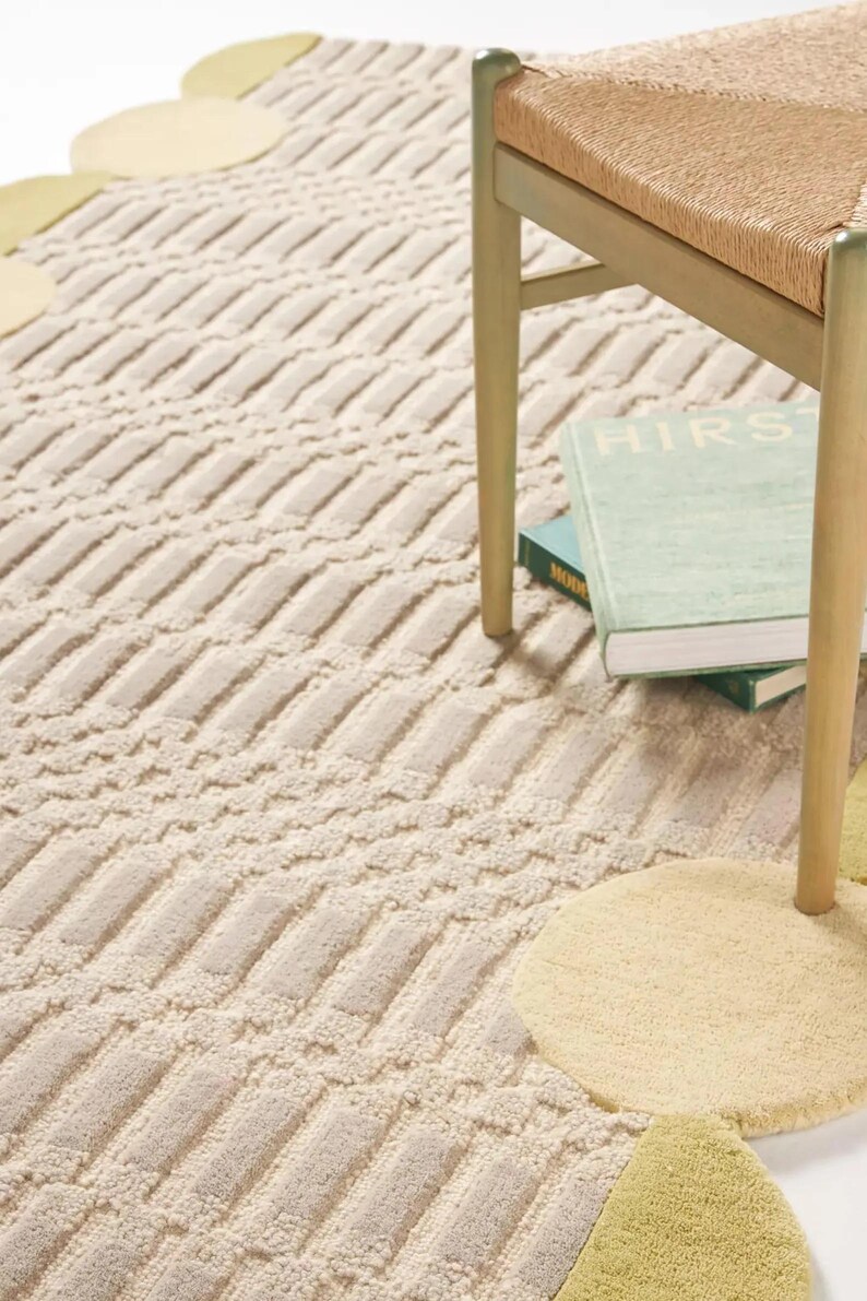 May include: Beige and cream-colored patterned rug with a geometric design. The rug has a textured surface and is paired with a wooden chair and books. The rug features decorative circular accents in yellow and green.