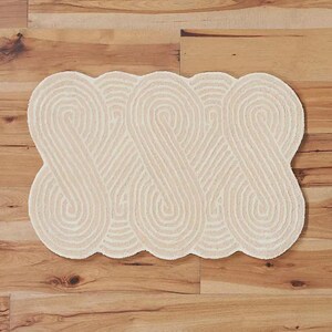 May include: Cream-colored, patterned rug with a repeating, intertwined design. The rug is rectangular with a scalloped edge and is placed on a wooden floor. The design features a series of connected, curved lines.