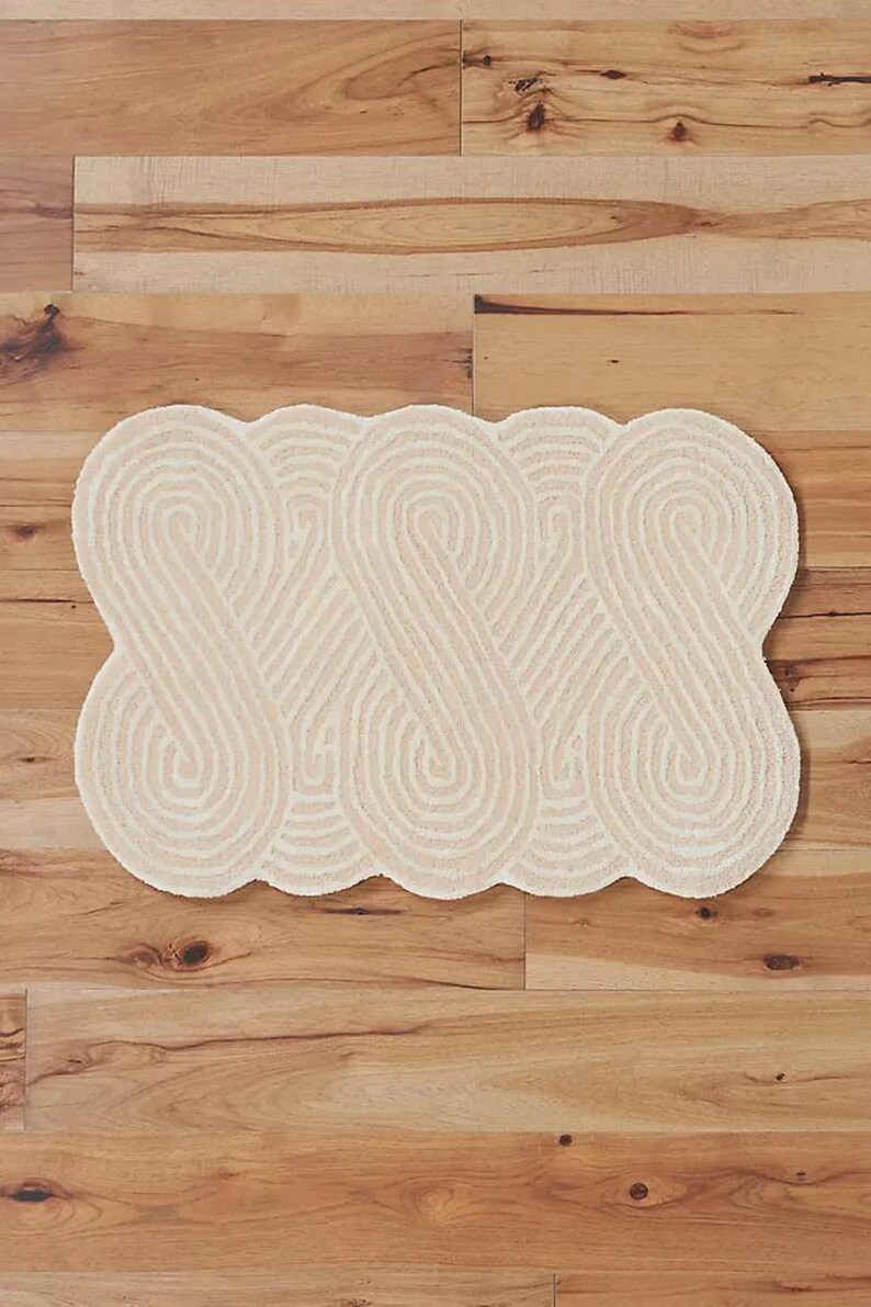May include: Cream-colored, patterned rug with a repeating, intertwined design. The rug is rectangular with a scalloped edge and is placed on a wooden floor. The design features a series of connected, curved lines.