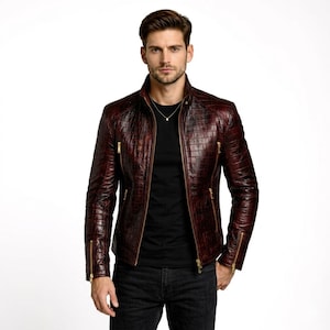 Può includere: Giacca in pelle bordeaux con motivo a rilievo effetto coccodrillo. La giacca presenta un collo alto, cerniere dorate e una vestibilità aderente. Il modello indossa una maglietta nera e jeans scuri.