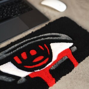 May include: Black rectangular rug with a stylized eye design. The eye features a black pupil, red accents, and a white iris, with a red tear-like shape extending from the eye. The rug is placed on a concrete surface, near a laptop and a white mouse.
