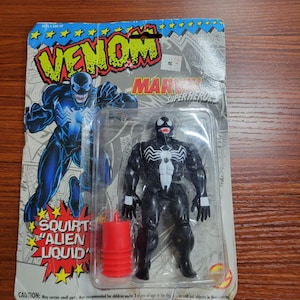 May include: A Venom action figure in original packaging featuring the text Venom, Marvel Super Heroes, and Squirts Alien Liquid. The black figure has a white spider emblem on the chest and white eyes. A red plastic accessory is included in the blister pack.
