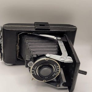 May include: A vintage black folding camera with a bellows mechanism, featuring a Kodet lens and Dak shutter. The lens assembly displays text including Kodet Lens, Dak Shutter, and aperture settings. A small Kodak logo is visible on the side panel.