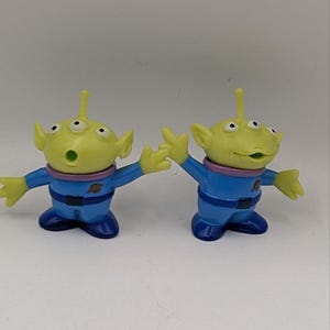 May include: Two small plastic alien figurines with lime green skin, three eyes, and antennae. They wear blue space suits with purple collars, dark blue belts, and dark blue boots. Each figure has a small planet emblem on the chest and open mouths.