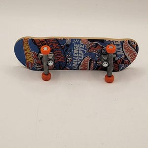 May include: A finger skateboard featuring a blue, black, and orange graphic design with the Hot Wheels logo and the text Challenge Accepted. The board has silver metal trucks and four small orange wheels attached to the underside of the deck.