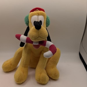 May include: A yellow plush dog toy featuring long black ears, wearing a red hat and green earmuffs. The plush character is holding a red and white striped candy cane in its mouth. The toy has a soft texture and is depicted in a sitting position.