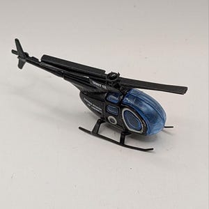 May include: A black toy helicopter with a translucent blue cockpit canopy. The body features the text "UNIT 12" and "UNITED STATES OF AMERICA" printed in white. It has a black main rotor, a tail rotor, and black landing skids.