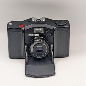 May include: A black Minox 35mm film camera with a fold-down front cover. The lens is labeled Color-Minotar 1:2.8 f=35mm. The camera body features a small red button on the top left, a central viewfinder, and a circular dial on the top right side.