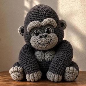 Könnte beinhalten: Handgehäkeltes Gorilla-Plüschtier in Grautönen. Das Gorilla hat ein lächelndes Gesicht, große Augen und detaillierte Häkelarbeiten. Das Plüschtier sitzt auf einer Holzoberfläche.