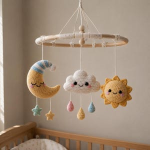 Könnte beinhalten: Handgefertigtes Baby-Mobile mit gehäkelter Sonne, Mond und Wolke. Die Sonne ist gelb mit einem lächelnden Gesicht, der Mond ist gelb mit einer blau-weiß gestreiften Mütze und die Wolke ist weiß mit rosa Wangen. Das Mobile enthält zwei Sterne und drei Tropfen.