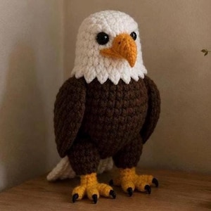 Könnte beinhalten: Eine handgefertigte Häkeladlerfigur. Der Adler hat einen weißen Kopf und Hals, einen braunen Körper und gelbe Füße mit schwarzen Krallen. Der Schnabel ist orange und die Augen sind schwarz. Der Adler steht auf einer Holzoberfläche.